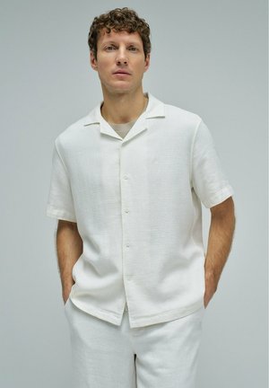 Hombre con pelo rizado que lleva una camisa blanca de manga corta abotonada y pantalones blancos a juego, de pie con las manos en los bolsillos.