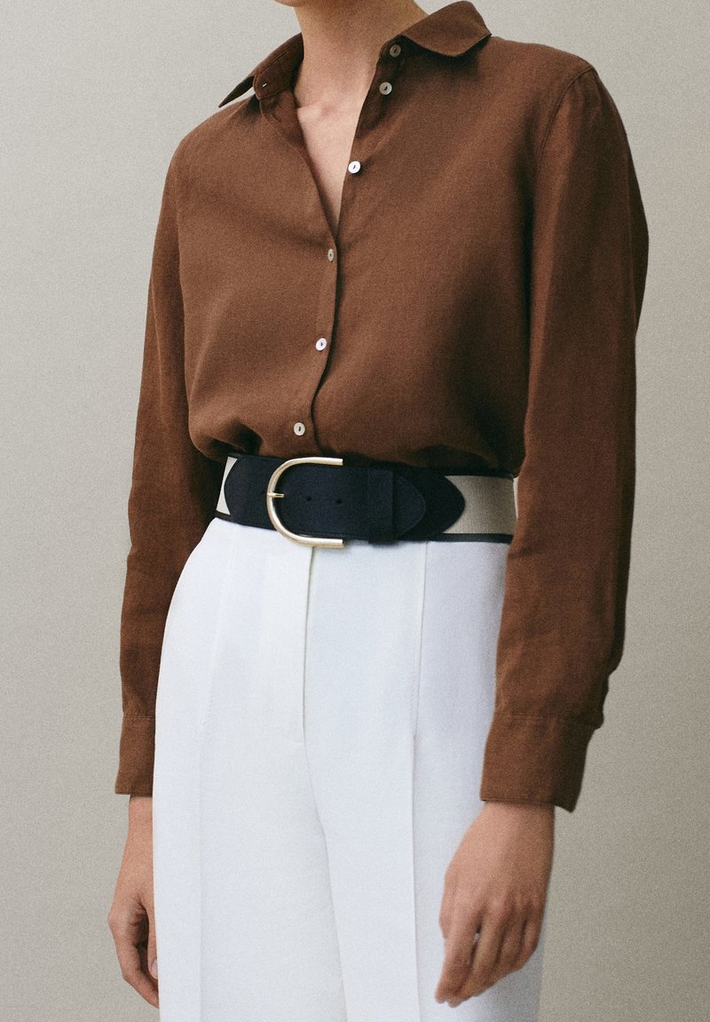 Personne portant une chemise marron boutonnée rentrée dans un pantalon blanc taille haute avec une large ceinture noire avec une boucle ovale dorée.
