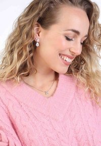 Silberne Ohrringe mit zwei klaren Edelsteinen, einem größeren tropfenförmigen und einem kleineren runden, getragen mit einem hellrosa, kabelstrickarmen Pullover.