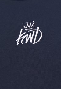 Vyšitá bílá koruna nad stylizovanými písmeny "KWID" na tmavě modrém pozadí.