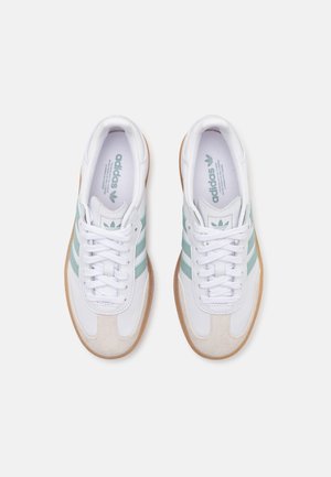 Paar weiße Adidas-Sneaker mit hellgrünen Streifen, Gummisohlen in Karamellfarbe und weißen Schnürsenkeln, von oben betrachtet.