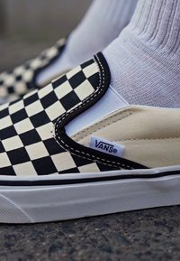 Svart og hvit sjakkbrett mønster lerret slip-on sneakers med kontrasterende svart søm og lavprofil hvit gummisåle. Har Vans-logo etikett.