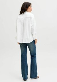 Hvid skjorte med knaplukning, en subtil prikmønster, lange ærmer og en buet søm, parret med blå udvidede jeans, set bagfra.