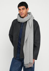 Casaco preto abotoado por cima de um suéter azul-marinho, combinado com um cachecol cinza claro e texturizado com franjas nas extremidades. Usado com jeans azuis.