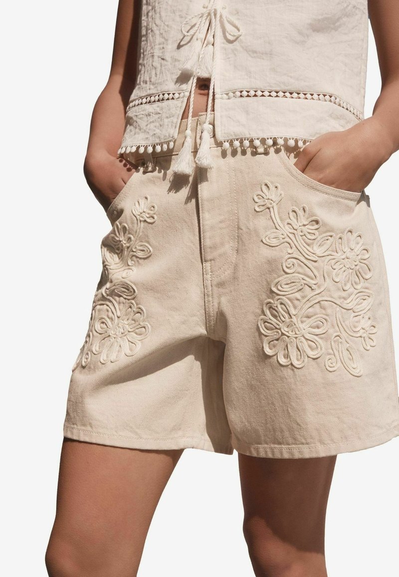 Shorts beiges brodés avec des motifs floraux sur les côtés, confectionnés en tissu texturé, dotés de poches et d'une coupe droite.