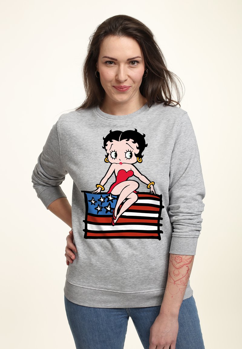 Sudadera gris que presenta un gráfico de una mujer de dibujos animados sentada sobre una bandera estadounidense. El diseño incluye colores rojo, azul y blanco con contornos negros.