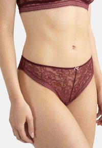 String en dentelle bordeaux avec des motifs floraux, des bords festonnés et un petit détail en ruban rose à la taille. Texture douce et transparente.