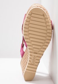 Sandale compensée avec semelle beige texturée, plateforme tressée et lanières roses, positionnée semelle vers le haut contre un mur blanc.