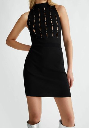 Černé mini šaty bez rukávů s vysokým límcem, vertikálními průstřihy ozdobenými malými korálky, nošené se stříbrnými kozačkami nad kolena.