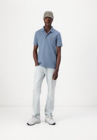 Lys blå polo skjorte med kortermet, gul logo på brystet, kombinert med lyse denim jeans og hvite sportssko. Grå caps.