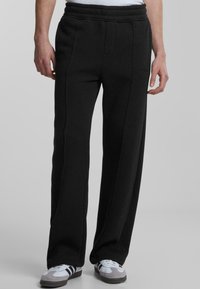 Black sweatpants with a relaxed fit, featuring a ribbed waistband, side seams, and smooth, textured fabric. Paired with grey and white sneakers.