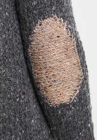 Maglione grigio lavorato a maglia con una superficie testurizzata, caratterizzato da una grande toppa beige sul gomito con un motivo a tessitura a contrasto.