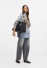 Γυναίκα που φοράει γκρι φούτερ και φαρδιά ξεθωριασμένα τζιν, κρατάει μια μεγάλη μαύρη τσάντα ώμου με γοητεία.