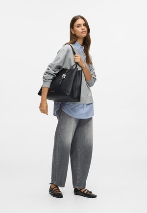 Kobieta w szarym swetrze i szerokich, wyblakłych dżinsach trzyma dużą czarną torbę na ramię, ozdobioną charmsami.