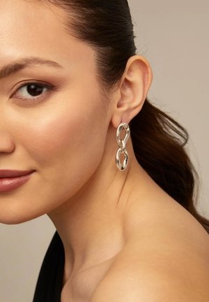 Boucles d'oreilles en argent avec un design ovale enchaîné. Surface lisse et brillante avec une forme moderne et sculpturale. Épaule et cou du modèle visibles.