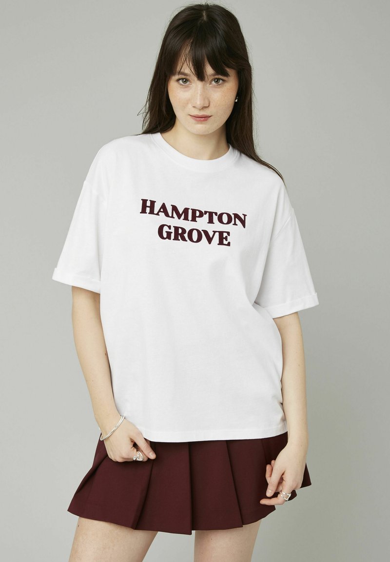 T-shirt en coton blanc avec "HAMPTON GROVE" en texte bordeaux, présentant une coupe décontractée et des manches courtes. Associé à une jupe plissée bordeaux.