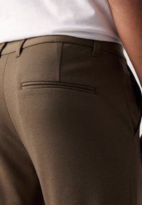 Pantalones de un verde oliva oscuro, confeccionados con una tela texturizada, que presentan un bolsillo trasero con una apertura estrecha y un corte ajustado.