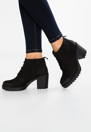 Femme portant des bottines noires à lacets avec talons épais et jean skinny foncé sur un fond blanc.