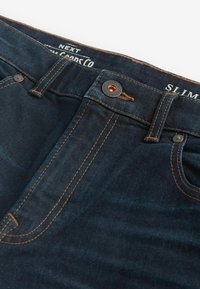 Jeans slim-fit de culoare albastru închis, din denim, cu cusături contrastante portocalii, un buton argintiu și o etichetă cu numele brandului pe talie.