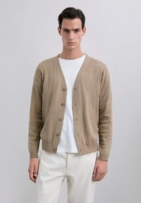 Cárdigan beige hecho de un material de punto suave, con un escote en V, cierre de botones y puños acanalados. Llevado sobre una camiseta blanca y pantalones claros.