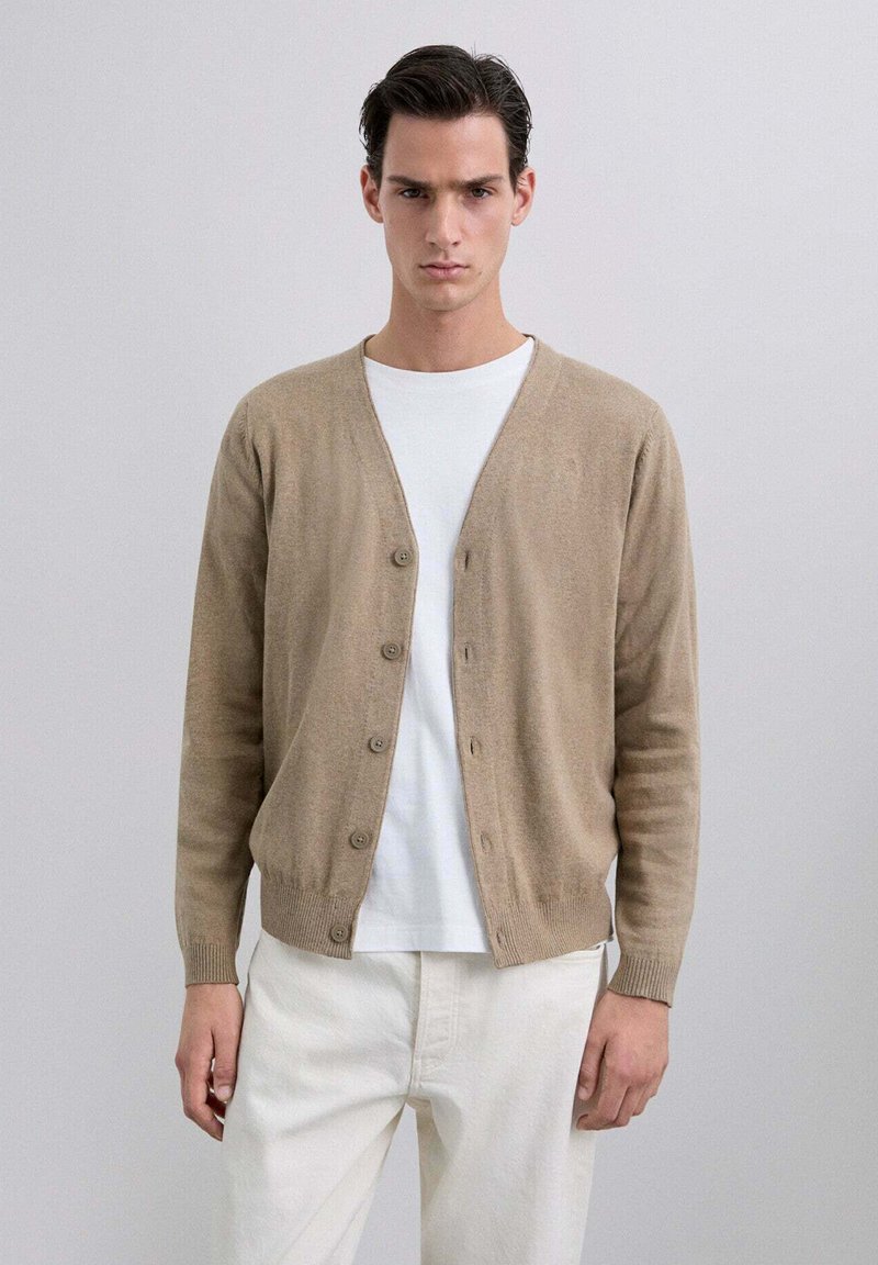 Cárdigan beige hecho de un material de punto suave, con un escote en V, cierre de botones y puños acanalados. Llevado sobre una camiseta blanca y pantalones claros.
