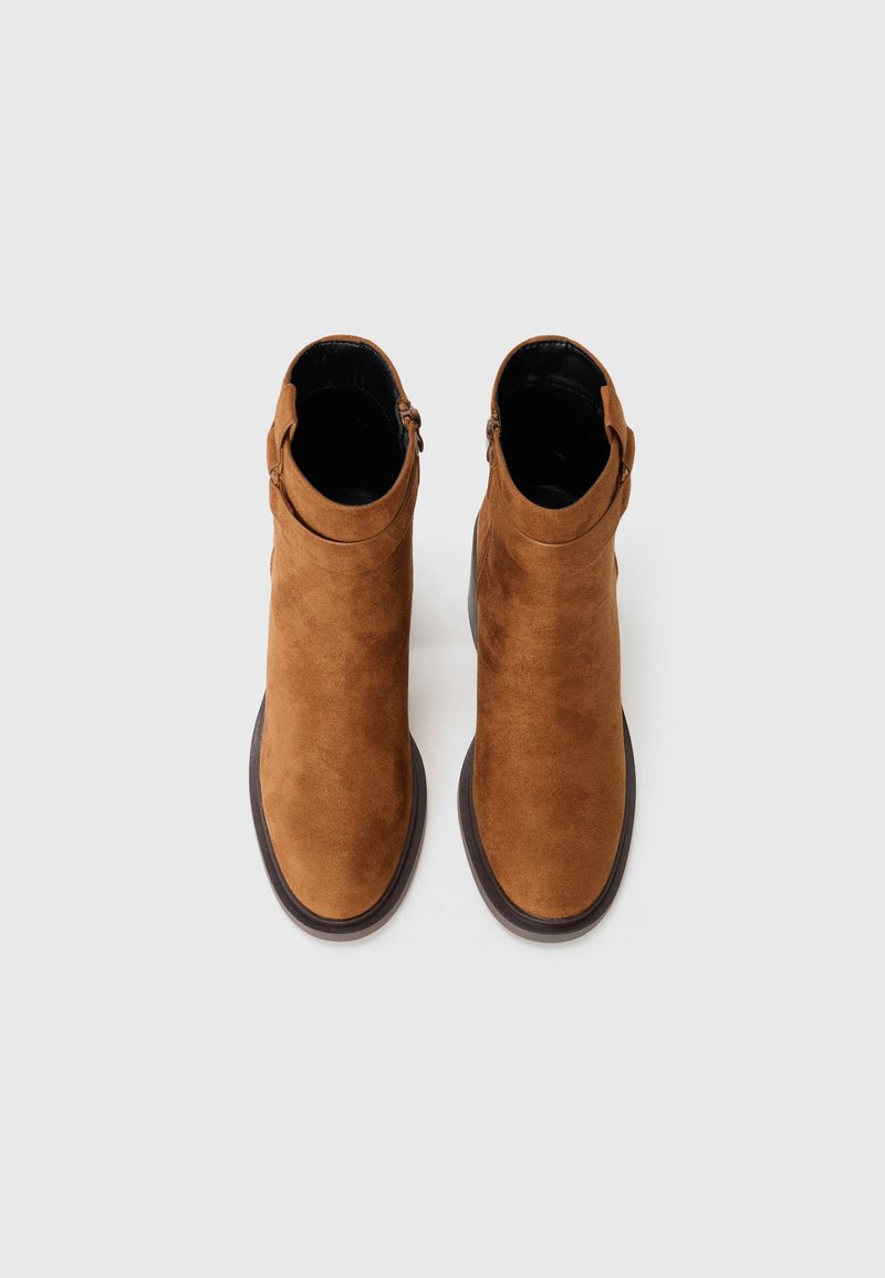 Botas ankle de ante marrón con una textura suave, puntera redondeada, un cierre lateral con cremallera y una suela de goma marrón en contraste.