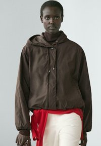 Frau trägt eine dunkelbraune Kapuzenjacke, einen burgunderroten Rollkragenpullover, weiße Hosen und einen roten Pullover, der um ihre Taille gebunden ist, und steht vor einem schlichten Hintergrund.