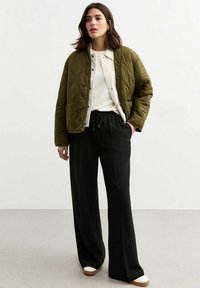 Veste bombardier matelassée kaki avec poches avant, superposée sur un haut beige, associée à un pantalon large noir et des chaussures claires.