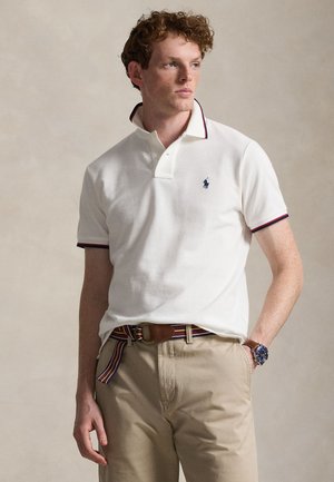 Mladić s kovrčavom kosom nosi bijelu polo majicu, bež hlače, prugasti remen i ručni sat, stoji s jednom rukom u džepu.