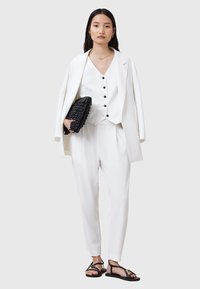 Completo bianco su misura con gilet abbottonato, pantaloni dal taglio morbido e sandali neri piatti. Tiene una pochette nera texturizzata. Accenti di gioielli semplici.