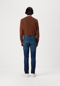 Ruskea pitkähihainen paita, jossa on pyöreä kaulus, yhdistettynä tummansinisiin slim-fit-farkkuihin ja valkoisiin lenkkareihin, nähtynä takaa.