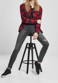 Ung person sidder på en sort skammel iført en rød ternet skjorte, sorte ripped skinny jeans og sorte høj-top sneakers.