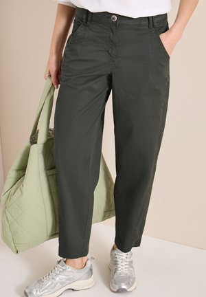 Frau trägt dunkelgrüne Hose und silberne Sneakers, hält eine hellgrüne gesteppten Tasche in der linken Hand, rechte Hand in der Tasche.