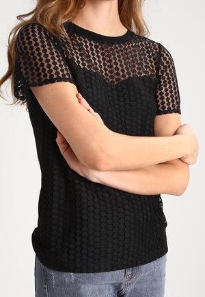 Kvinde iført en sort kortærmet top med et gennemsigtigt prikmønster over brystet og ærmerne, kombineret med blå jeans.