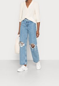 Lys cremefarvet bluse med knaplukning og blonderkant, kombineret med lyseblå, slidte, vidde-bukser, hvide sneakers og en beige håndtaske.