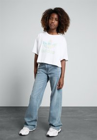 T-shirt branca cropped com o logótipo iridescente da Adidas, combinada com jeans de perna larga azul claro e ténis brancos com detalhes pretos.