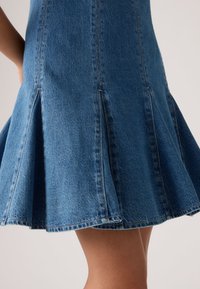 Μπλε τζιν φούστα καμπάνα με ραμμένες λεπτομέρειες πάνελ, φορεμένη από άτομο, ορατή από τη μέση μέχρι το μέσο του μηρού, με ουδέτερο φόντο.