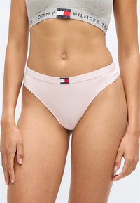 Femme portant un sous-vêtement rose clair de Tommy Hilfiger et un crop top gris assorti avec une bande logo Tommy Hilfiger, debout avec les bras relâchés.