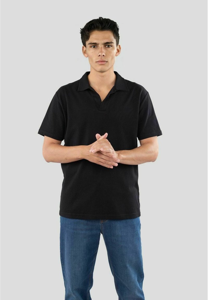 Sort polo-shirt med en afslappet pasform, korte ærmer og spids krave, parret med blå jeans. Modellen står med hænderne foldet.