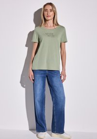 T-shirt à manches courtes vert clair avec "NEVER DOUBT" imprimé en noir. Porté avec un jean bleu à jambe large et des chaussures blanches. Style décontracté.