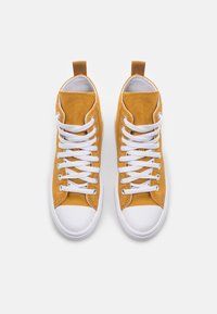 Zapatillas de lona amarillas de caña alta con cordones blancos, punteras de goma blanca y una pestaña de tela en el talón para facilitar su uso.