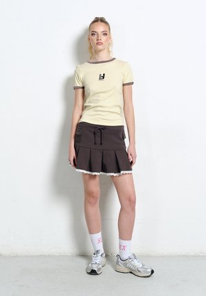 Młoda kobieta stoi na tle białej ściany, ubrana w kremową bluzkę z krótkim rękawem, ciemną plisowaną spódnicę z kieszeniami, białe skarpetki i srebrne trampki.