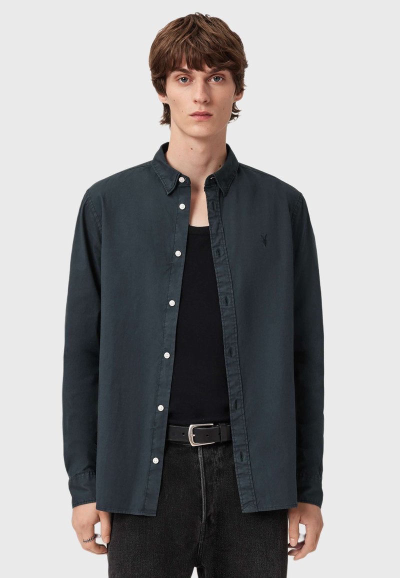 Camicia button-up di un blu navy scuro in tessuto morbido, con colletto classico, bottoni bianchi e un delicato dettaglio del logo sul petto. Indossata sopra un top nero.