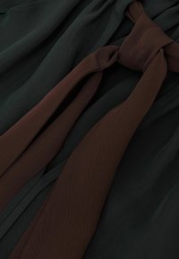 Bruine stoffen stropdas met een losse knoop over donkergroen gestructureerd stof, met gedetailleerde textuur en plooien van beide materialen.