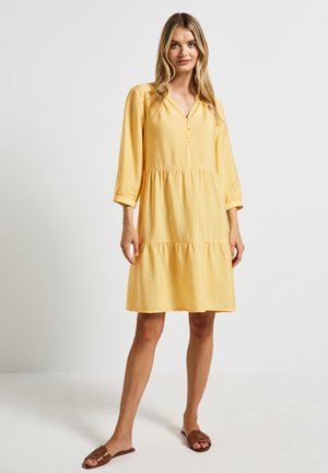 Abito giallo lungo fino al ginocchio in tessuto leggero con un design a strati, caratterizzato da un décolleté a V e maniche a trequarti, abbinato a sandali marroni.