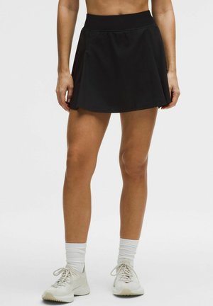 Femme portant une jupe de sport noire, des chaussettes blanches et des baskets blanches, debout les jambes légèrement écartées sur un fond uni.