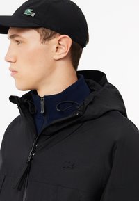 Giacca nera con cappuccio e colletto con zip, caratterizzata da uno strato interno navy testurizzato e un logo nero ricamato. Il cappuccio è anch'esso nero.