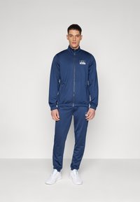 Navy tracksuit made of smooth fabric with a zip-up jacket and elastic cuffs; features logo on the left chest and side zipper pockets.
