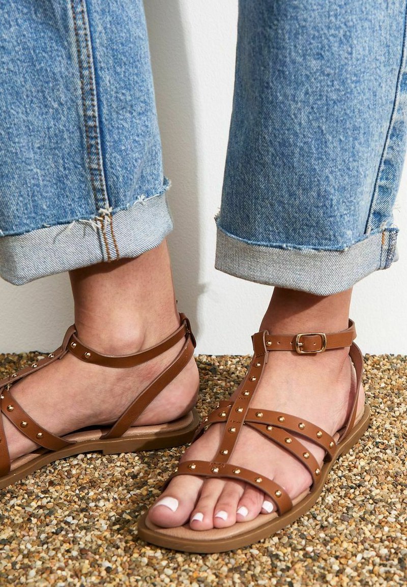 Bruna remsandaler med flera band och metallspikar, platt sula och justerbar ankelsn buckle, bärs med uppvikta blå jeans.