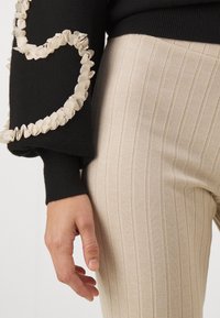 Jersey negro con volantes de ribete crema en las mangas, combinado con pantalones beige acanalados con rayas verticales. Mano apoyada en el pantalón.